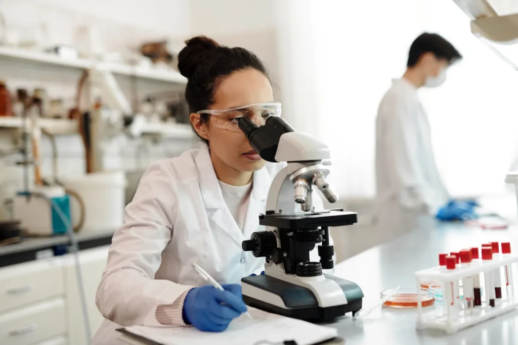 A researcher in a laboratory looks through a microscope while taking notes, with sample tubes on the bench