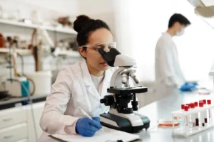 A researcher in a laboratory looks through a microscope while taking notes, with sample tubes on the bench