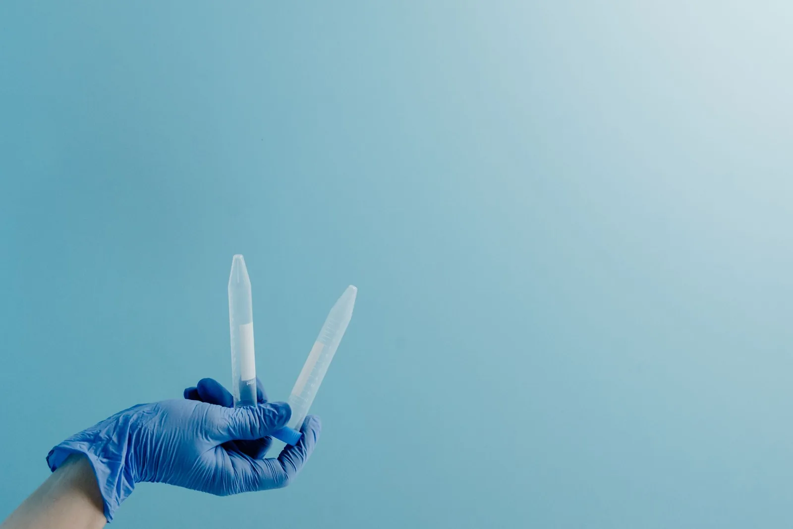 A gloved hand holds two laboratory sample tubes against a soft blue background, representing a genetic test sample