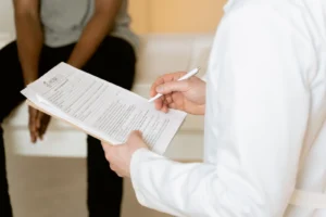 A clinician in a white coat reviews a form with a seated patient during a consultation about genetic testing
