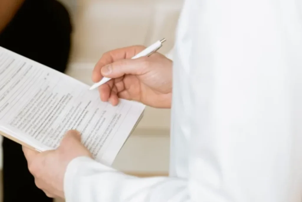 Close view of a clinician in a white coat writing on a consultation form, with the patient seated beyond
