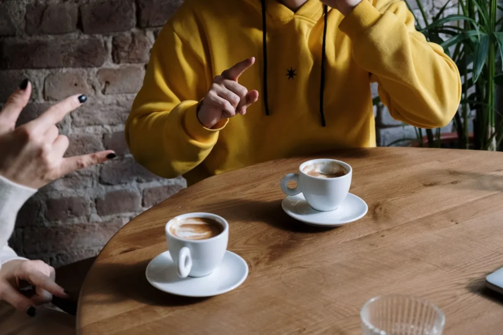 Two people sit at a cafe table with coffee and talk to each other in sign language